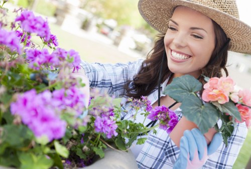 A woman with a cheerful expression, wearing a straw hat and light blue gloves, tending to a garden filled with vibrant purple, pink, and peach flowers. She is in an outdoor garden space with visible greenery and sunlight, using her hands to carefully handle the flowering plants. The garden features a variety of foliage and flowering plants, with some showing lush green leaves, and others displaying colorful blooms. The scene suggests regular gardening activity aimed at maintaining a healthy and aesthetically pleasing outdoor environment. The background slightly blurs out trees and a lawn, indicating a well-maintained garden area typical of residential landscaping in East Ham. This image highlights professional gardening care, including planting, tending, and garden beautification, suitable for a landscaping service website focused on garden renovations and outdoor landscaping improvements for local homes and gardens.