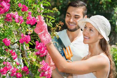 A woman and a man are in a lush garden, tending to a vibrant flowering shrub with bright pink blossoms. The woman, wearing a wide-brimmed straw hat and gardening gloves, is actively pruning the bush with a small hand pruner, while the man, dressed casually, stands close by holding a gardening fork. The garden features a neatly maintained lawn in the foreground, with the flowering shrub taking prominence on the left side of the scene. Behind them, there are tall green trees and other shrubs that create a natural backdrop, suggesting a well-tended outdoor space typical of East Ham gardens. The lighting indicates a sunny day with clear weather, enhancing the natural tones of the green foliage and colorful flowers. Visible soil areas and a section of paved pathway near the plants indicate landscape features that can be maintained through professional gardening services from Landscapers East Ham. This scene exemplifies typical garden maintenance activities involved in garden renovation and enhancement projects, aligning with local gardening and landscaping practices in London postcodes around E6 and nearby areas.