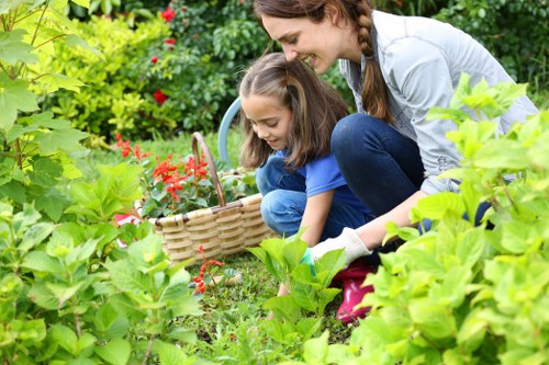 A woman and a young girl are engaged in gardening in a well-maintained outdoor space, surrounded by lush green foliage, vibrant red flowering plants, and diverse shrubbery. The woman is kneeling on the grass, leaning forward with a smile, while the girl is holding a wicker basket filled with small plants or flowers, seemingly preparing for planting. The garden features a neatly bordered flower bed with dense, healthy grass and a variety of leafy plants and shrubs, creating a colorful and inviting environment. In the background, there are more plants and a backdrop of trees, indicating a mature and landscaped garden typical of East Ham. The scene suggests outdoor maintenance and planting activities, reflecting professional gardening services offered by Landscapers East Ham, with natural light highlighting the vivid greens and reds, and a clean, organized layout suitable for a residential garden in the London area.