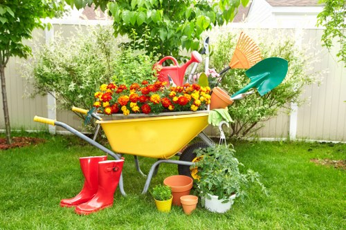 A well-maintained residential garden featuring a lush green lawn with evenly trimmed grass, bordered by a white wooden fence and surrounded by various shrubs and small trees. In the foreground, there is a yellow plastic wheelbarrow filled with vibrant red and yellow flowering plants, placed on the grass. Nearby, a pair of red rubber gardening boots rest next to several empty flower pots of different sizes, some terracotta and some plastic. A plastic watering can is positioned behind the wheelbarrow, along with gardening tools and a green garden trowel. A blue plastic shovel and an orange leaf rake are leaning against the wheelbarrow, with a white potted plant with leafy foliage situated next to them. The background includes more greenery, with a clear sky and natural daylight enhancing the scene, indicative of spring or summer. This outdoor space aligns with quality lawn and garden maintenance services offered by Landscapers East Ham in the East Ham area, supporting local gardening and landscaping needs.