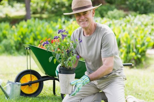 A mature man is kneeling outdoors in a well-maintained garden in East Ham, holding a potted plant with purple and red flowers, likely to be added to a flower bed or garden border. He is dressed in light gardening clothes, including a wide-brimmed hat, grey t-shirt, and gardening gloves. Behind him, a green wheelbarrow with a black and yellow wheel is positioned on lush, neatly mown grass, and a watering can rests on the ground nearby. The garden features a dense backdrop of greenery, with hedgerows or shrubs, and mature trees providing shade. The soil in the flower bed appears dark and healthy, bordered with natural stones or edging materials. The outdoor scene is bathed in natural daylight, suggesting a bright, calm day suitable for gardening work. This image captures the typical outdoor environment managed by a professional gardening service like Landscapers East Ham, emphasizing landscaping and plant care activities in a suburban garden setting.

Note: The garden layout includes a grass lawn in the foreground, a wheelbarrow with gardening tools, and planting elements, all contributing to a neat, organized outdoor space characteristic of the East Ham area.
