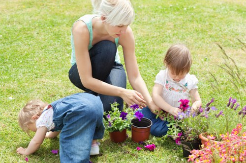 A woman with short blonde hair, dressed in a sleeveless light blue top and dark jeans, is kneeling on a well-maintained grassy lawn in a garden area with two young children, a girl and a boy. The woman is tending to potted plants and flowers, which include vibrant purple, pink, and purple blooms, some in black plastic pots and others directly in the ground. The children, dressed in casual clothing; the girl in a white top with pink accents and the boy in a light blue outfit, are actively exploring and interacting with the garden plants. The lawn surface is lush and evenly cut, with a backdrop of additional flower beds and garden greenery. The scene is set outdoors on a bright, slightly overcast day, with natural light illuminating the vibrant colours of the flowers and greenery, illustrating a typical seaside garden environment near East Ham, suitable for outdoor gardening activities supported by professional landscaping services such as those from Landscapers East Ham.