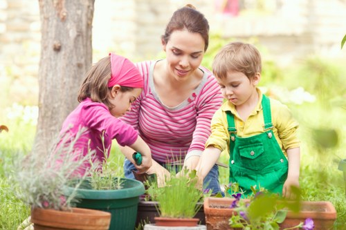A woman with dark hair pulled back, wearing a pink and white striped long-sleeve top, is engaged in gardening activities in a well-maintained outdoor garden space. She is closely supervising and assisting two young children, a girl wearing a pink headband and a purple top, and a boy in a yellow shirt with green overalls, as they plant in various pots and flower beds. The garden features a lush, green lawn area, bordered by a mature tree with textured bark, and is surrounded by a low brick wall. The scene showcases healthy, vibrant plants with green foliage, some with purple flowers, and fresh soil in rectangular terracotta planters and plastic pots. The background includes additional greenery, suggesting a landscaped backyard environment, possibly part of a residential property in East Ham. The weather appears sunny and mild, with natural daylight illuminating the scene, highlighting the engaging outdoor activity focused on gardening and outdoor upkeep, consistent with landscaping services provided by Landscapers East Ham.